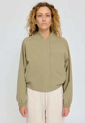 Woman with curly blonde hair wears a light olive green jacket and cream-colored pants, standing against a plain light background.