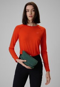 Femme aux cheveux foncés mi-longs portant une chemise orange à manches longues et un pantalon noir, tenant une veste verte pliée à hauteur de taille.