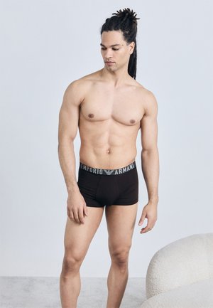 Uomo senza maglietta con dreadlocks che indossa boxer neri Emporio Armani, in piedi accanto a una sedia bianca dalla texture, su uno sfondo semplice.