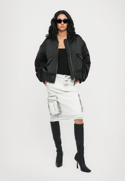 Veste bomber en satin noir, débardeur noir, jupe en denim blanche avec poches, et bottes noires à talons aiguilles jusqu'aux genoux.