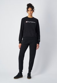Felpa nera in cotone con logo "Champion" bianco, scollo rotondo, polsini a costine, abbinata a pantaloni jogger neri e sneakers nere.