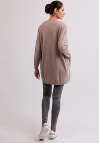 Lange, lichtbruine sweater met een relaxte pasvorm, zijzakken en ribbelzoom, gecombineerd met grijze skinny jeans en witte sneakers.