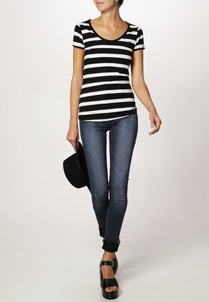 Woman walking wearing black and white striped t-shirt, blue rolled-up skinny jeans, black platform heels, holding a black hat in left hand.