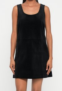 Femme portant une mini-robe noire en velours côtelé sans manches, avec un col rond et un ourlet droit, debout devant un fond uni.