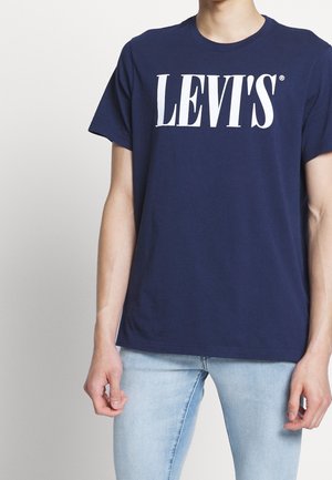 Osoba ubrana w granatowy t-shirt Levi's i jasnoniebieskie dżinsy, stojąca na jednolitym, jasnoszarym tle.