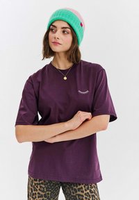 Fialové bavlněné tričko s krátkými rukávy a bílou výšivkou ve tvaru řasy, spojené s kalhotami s leopardím potiskem, a doplněné barevnou čepicí.
