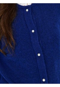 Modra pletena jopa z teksturirano površino, okrašena z okroglimi bisernimi gumbi spredaj. Dolgi rokavi in zaobljen izrez na vratu.