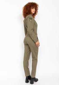 Mono caqui con mangas largas, bolsillos delanteros, detalles de estilo militar y un diseño ajustado. Combinado con botines negros de tacón.