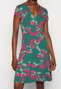 Kortärmad, grön klänning med v-ringning, prydd med ett blommönster i rosa och ljusgrönt, tillverkad av mjukt, texturerat tyg.