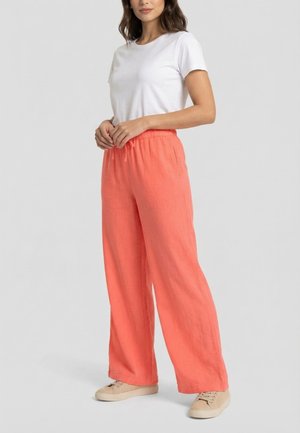 Mujer con camiseta blanca de manga corta, pantalones amplios color coral con cintura elástica y zapatillas casuales beige, de pie frente a un fondo liso.