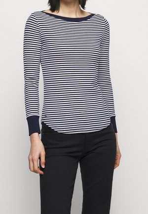 Femme portant une chemise à manches longues à rayures horizontales bleu marine et blanches avec des poignets bleu marine, et un pantalon noir, debout devant un fond uni.