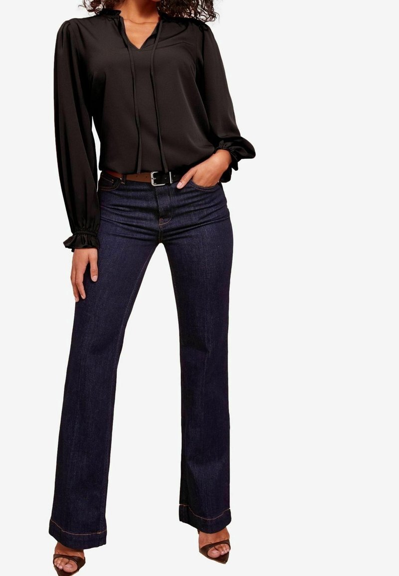 Femme portant un chemisier noir à manches longues, un jean bootcut bleu foncé avec une ceinture marron, et des talons ouverts marron, debout avec une main dans la poche.