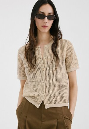 Mujer con cárdigan beige de manga corta y punto abierto con botones, pantalones plisados verde oliva y gafas de sol pequeñas ovaladas negras.