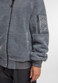 Giacca in pile grigia con zip frontale, polsini a coste e corpo testurizzato. Presenta una tasca sul braccio destro con chiusura a zip e dettaglio con bottone a pressione.