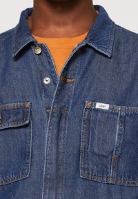 Chemise en denim bleu foncé avec col classique, deux poches poitrine, sous-vêtement orange contrastant et boutons logo en métal.