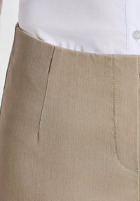 Beige hoge taille broeken gedragen met een ingestopt wit shirt, met zichtbare stoftextuur en stikdetails bij de taille.