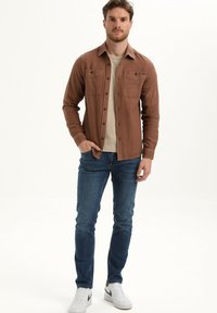 Rjava srajca z dolgimi rokavi in gumbom na sprednji strani, z dvema žepoma na prsih, v kombinaciji z modrimi kavbojkami in belimi športnimi copati. Preprost dizajn in tkanina iz prepletenih vlaken.