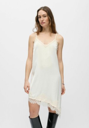 Ženska, oblečena v brezrokavno belo obleko z aplikacijo iz čipke in črne škornje do kolen, stoji pred preprostim svetlim ozadjem.