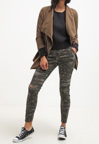 Brązowa kurtka z długim rękawem noszona na czarnym swetrze, w połączeniu z wąskimi spodniami w kamuflażu z przetarciami i czarnymi trampkami.