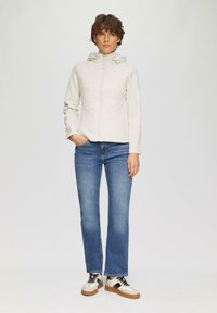 Veste à capuche beige clair avec texture matelassée, fermeture éclair devant et deux poches latérales, associée à un jean en denim bleu et des chaussures décontractées.