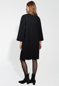 Zwarte oversized jas met lange mouwen, een gladde textuur, rechte snit en minimale details, gecombineerd met zwarte enkellaarsjes en doorschijnende panty's.