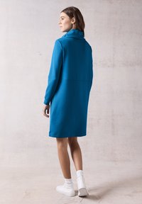 Vihreä-sininen huppari-mekko, jossa on korkea kaulus ja pitkät hihat. Mekossa on rento malli ja hienovarainen ommel vyötäröllä.