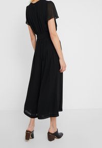 Vestido negro plisado con mangas cortas y transparentes, cintura ceñida y silueta fluida, combinado con mule negros de tacón.