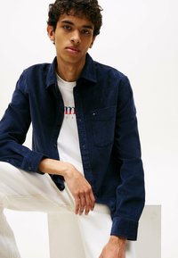 Junger Mann in einem dunkelblauen Cordhemd über einem weißen T-Shirt mit Grafik, der mit einem Arm auf seinem Knie sitzt, vor einem weißen Hintergrund.