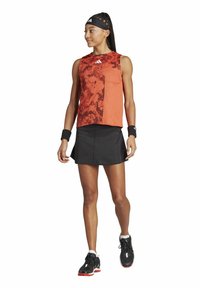 La indumentaria de tenis incluye una camiseta sin mangas de color rojo con un diseño estampado y una falda negra. Los accesorios incluyen muñequeras y zapatillas de tenis negras.