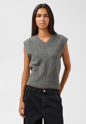 Jeune femme aux longs cheveux foncés portant un pull sans manches gris en tricot et un pantalon noir taille haute sur un fond uni.