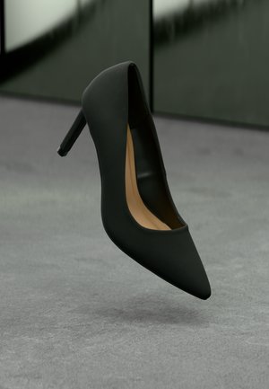 Sort spids tå højhælet sko med stiletthæl, beige inderside, placeret oprejst på grå overflade med sløret grøn baggrund.