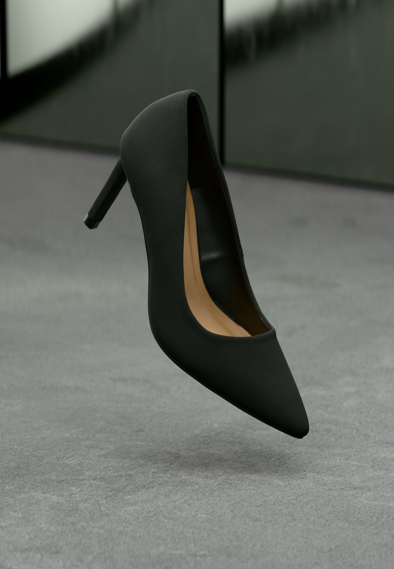 Escarpin noir à bout pointu avec talon stiletto, intérieur beige, positionné debout sur une surface grise avec un arrière-plan vert flou.
