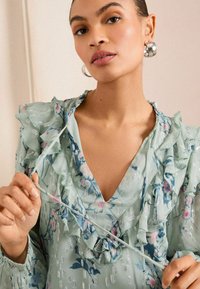 Gėlėmis marginta, šviesiai žalia bluzonė su raukiniais aplink iškirptę ir rankoves, turinti V formos iškirptę ir tekstūruotą medžiagą su subtiliu blizgesiu.