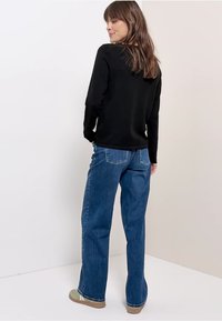 Frau mit braunen Haaren, trägt ein schwarzes Langarmshirt, blaue Jeans mit geradem Bein und beige Schuhe, steht vor einem schlichten hellen Hintergrund.