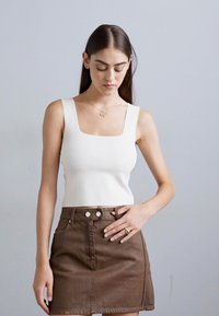 Pikslite naine, kellel on pikad tumepruunid juuksed, kannab valget ruudukujulise kaelusega ilma varrukateta toppi ja pruuni nahast mini seelikut, vaatab allapoole, hoides ühe käega seeliku peal.