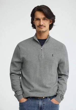 Hombre con cabello oscuro de longitud media y vello facial claro, vestido con un suéter gris con cremallera hasta el pecho y vaqueros azules, manos en los bolsillos, fondo neutro.