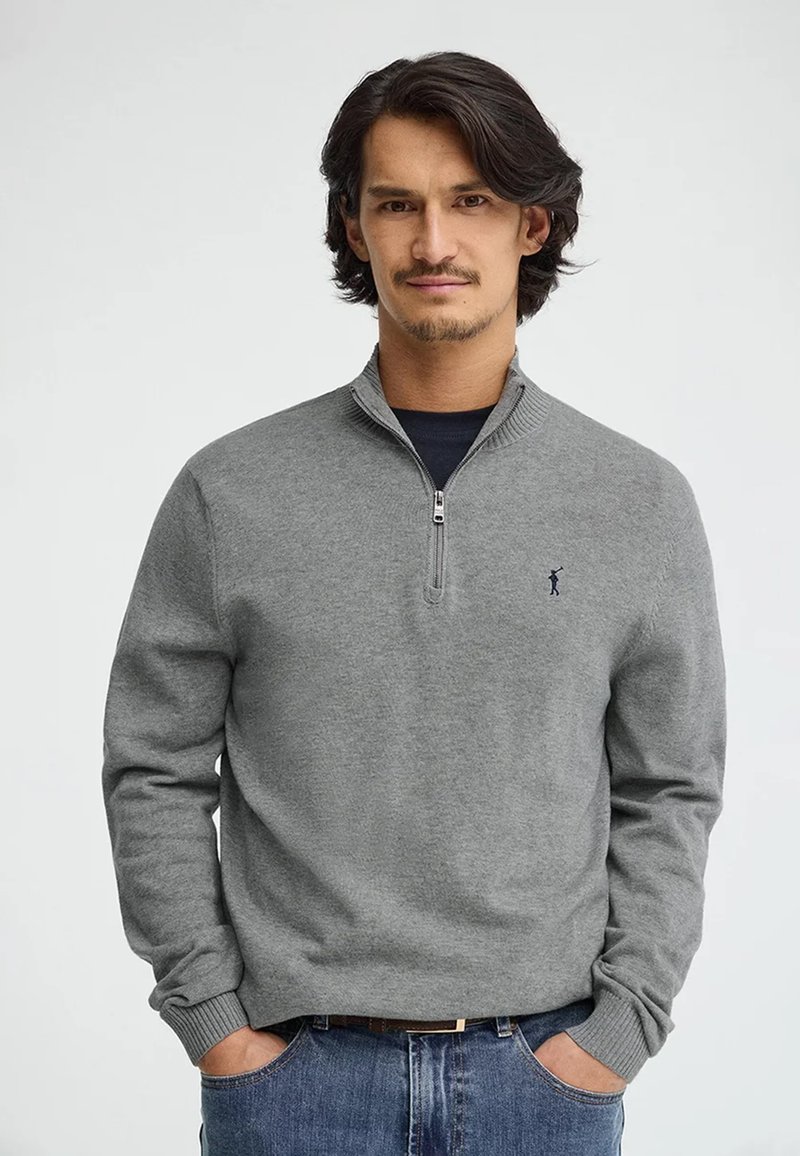 Mann mit mittellangem dunklem Haar und leichtem Gesichtshaar, der einen grauen Pullover mit Viertel-Reißverschluss und blaue Jeans trägt, Hände in den Taschen, neutraler Hintergrund.