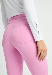 Rosa stretchbyxor med bakfickor, med kontrasterande sömmar och varumärket "Röhnisch" präglat på midjebandet. Slät textur.