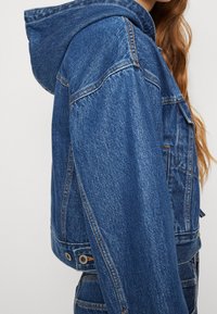 Modrá džínová bunda s kapucí, oříznutá, s viditelným prošíváním, knoflíkovými detaily a strukturovanou tkaninou. Boční pohled ukazuje design.