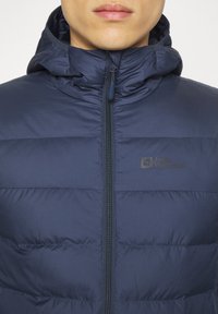 Giubbotto imbottito blu navy con cappuccio, caratterizzato da sezioni trapuntate, chiusura con zip completa e un logo sulla parte del petto. Texture liscia.