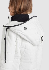 Veste matelassée blanche avec un col noir, dotée d'une fermeture éclair et de boutons, et ornée de tissus texturés et de coutures contrastantes.