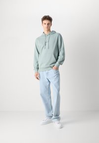 Felpa verde chiaro con tasca frontale e cordoncino, abbinata a jeans dritti azzurri e sneakers bianche. Design semplice, vestibilità rilassata.