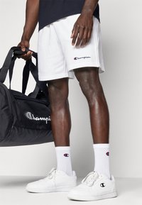 White athletic shorts with embroidered logo, paired with white socks featuring logo, white sneakers, and a black duffel bag.