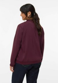 Burgunderfarbene Jacke mit langen Ärmeln, rundem Ausschnitt und gerippten Bündchen. Glattes Material mit lockerer Passform, kombiniert mit dunklen Jeans.