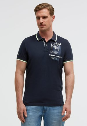 Mann trägt ein marineblaues Poloshirt mit weißem und grün abgesetztem Kragen und Ärmeln, bedrucktem Text und Palmenmotiv auf der Brust, kombiniert mit hellblauen Jeans.