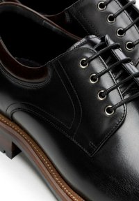 Czarne skórzane buty o gładkiej powierzchni, wyposażone w sznurowane zapięcie, metalowe przelotki oraz drewnianą podeszwę z jasnobrązowym akcentem.