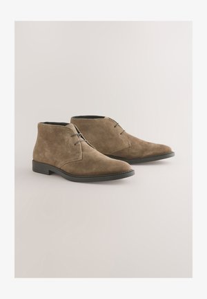 Braune Wildleder-Stiefeletten mit glatter Textur, ausgestattet mit niedrigem Absatz, rundem Zehenbereich und Schnürsenkeln. Dezente Nahtdetails in der Nähe der Zunge.