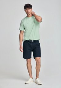 Ljusgrön bomull t-shirt med subtilt logo, parat med mörkblå shorts och vita sneakers. Avslappnad passform med korta ärmar och bältesdetalj.