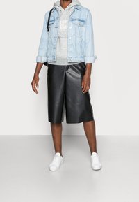 Persona che indossa una giacca di denim blu chiaro sopra un cappuccio grigio, pantaloni in pelle neri fino al ginocchio, sneaker bianche e porta una borsa a tracolla nera.