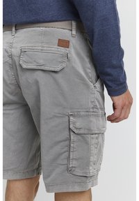 Light gray cargo shorts in cotton blend, featuring side and back pockets, adjustable waistband, and textured fabric. Leather brand patch included.